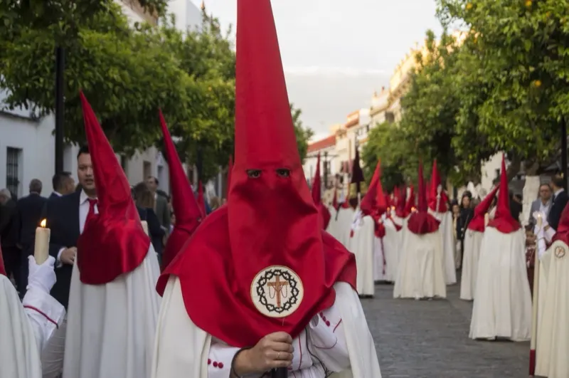 Nazareno de capirote rojo con el escudo de la Hermandad, cirio en mano