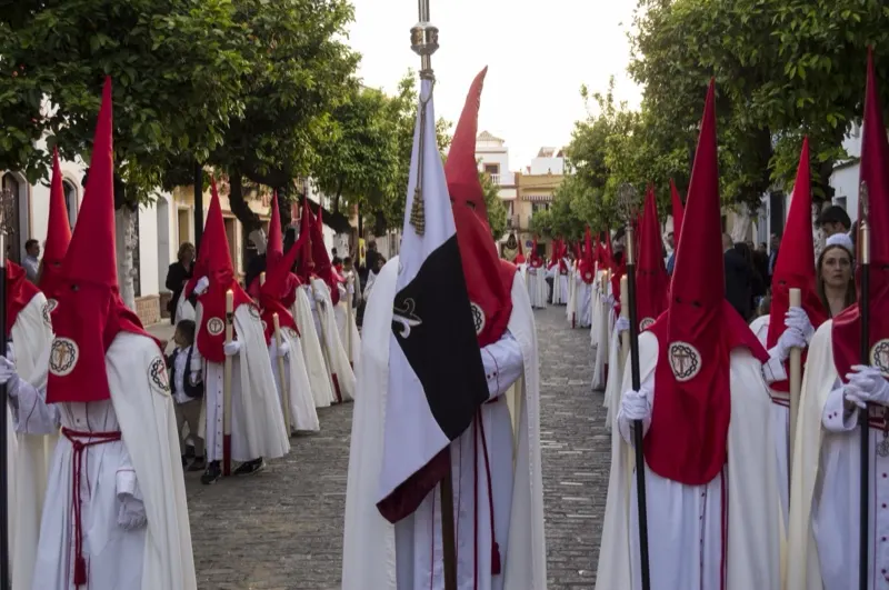 Tramo de nazarenos de túnica blanca y capirote rojo, con la bandera de la Hermandad